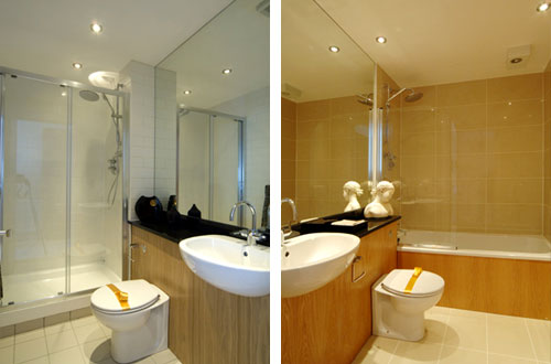 Nowoczesna łazienka z kabiną prysznicową, umywalką wpuszczoną w drewnopodobny blat i toaletą z żółtą taśmą zabezpieczającą, odbicie w lustrze ukazuje wannę z parawanem.